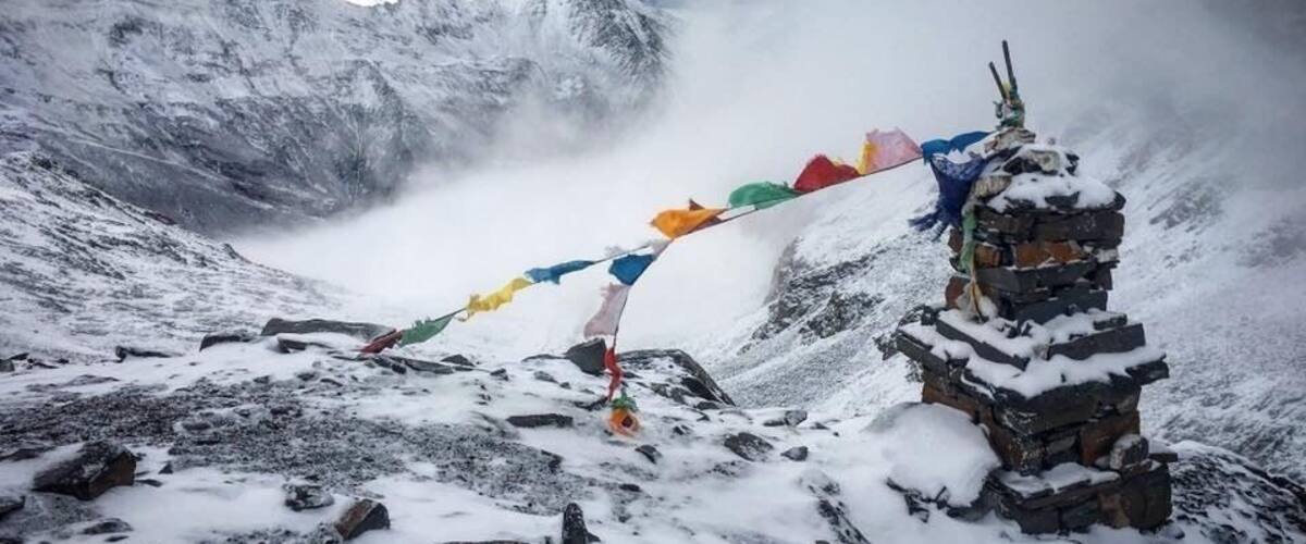 A snow-covered mountain peak with a small shrine and colorful prayer flags.