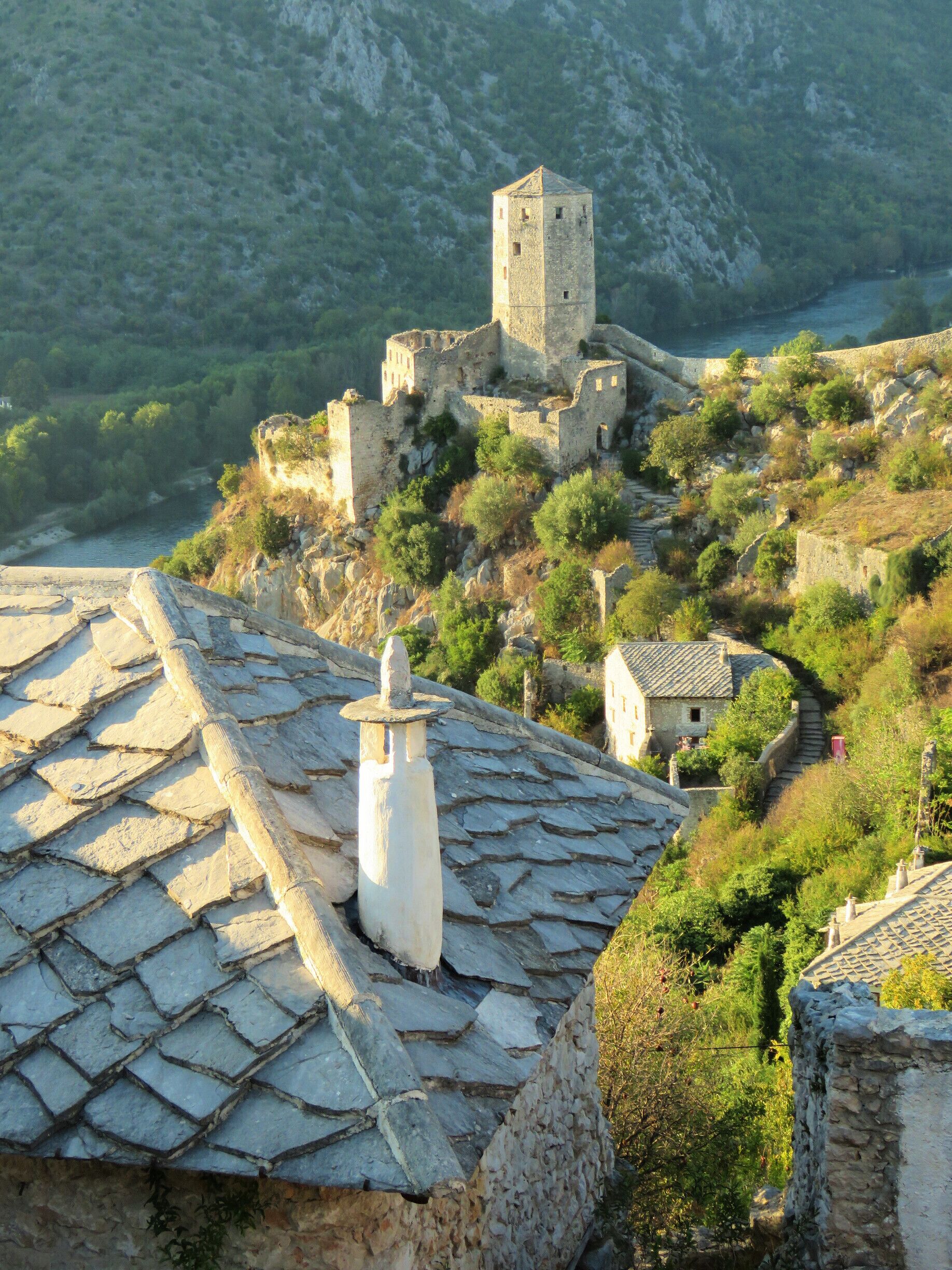A historic stone fortress with a tower, surrounded by lush greenery and a river.