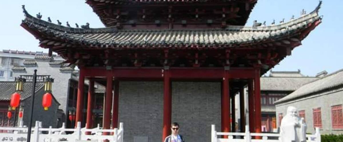 A traditional Chinese pavilion with a tiled roof and red pillars, a statue, and a person standing in front.