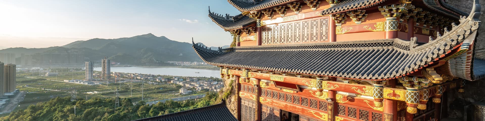 A traditional Chinese temple with ornate architecture, a stone lion statue, and a view of a cityscape and mountains in the background.