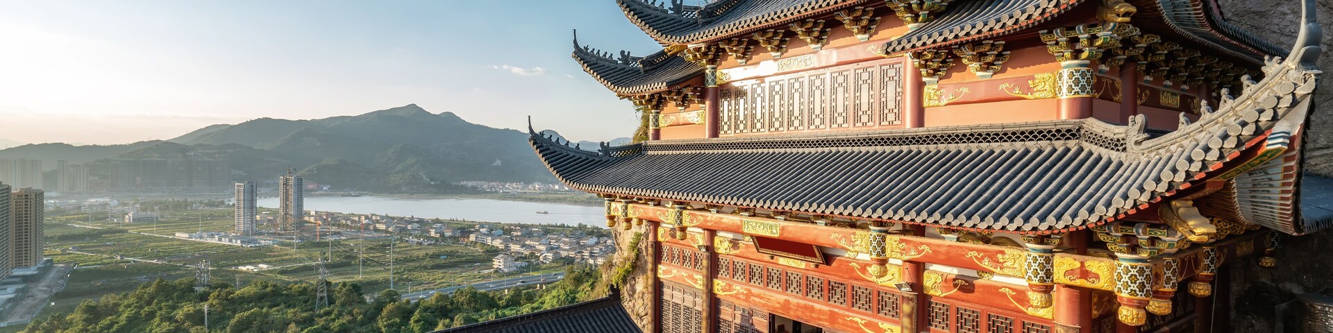 Um templo tradicional chinês com arquitetura elaborada, uma estátua de leão de pedra e vista para a paisagem urbana e montanhas ao fundo.