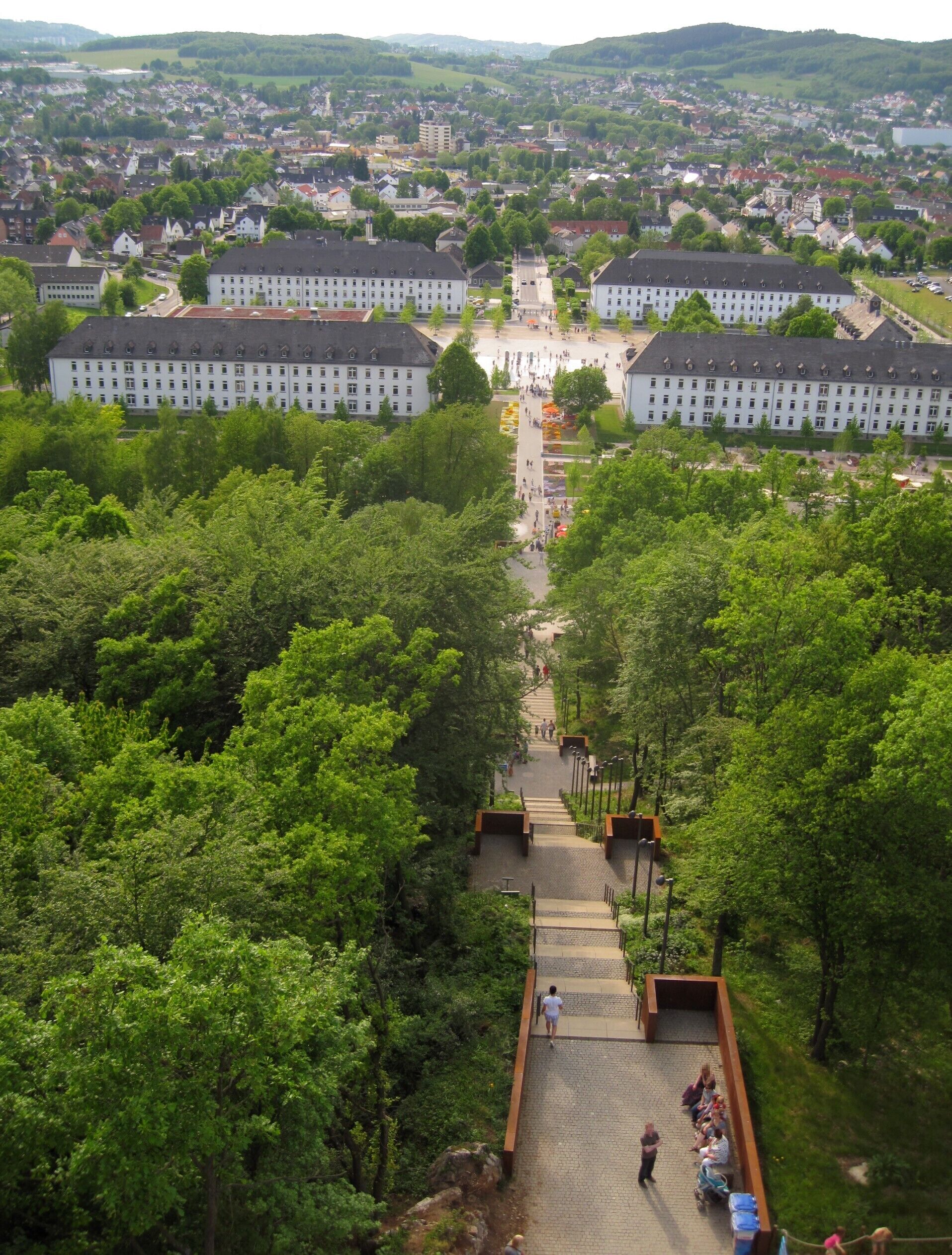 A town with a central square surrounded by lush green trees and a large white building with a clock tower.