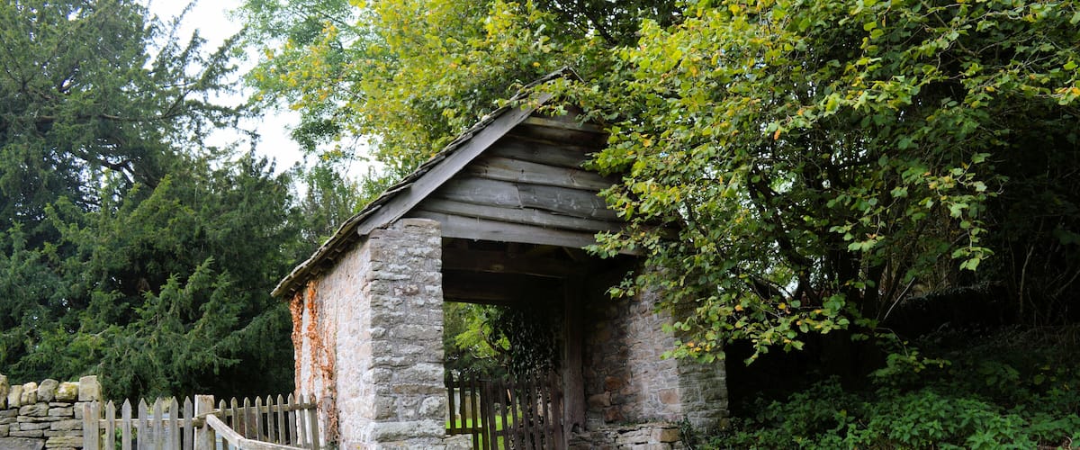 A rustic stone structure with a wooden roof, surrounded by lush greenery and a stone fence.
