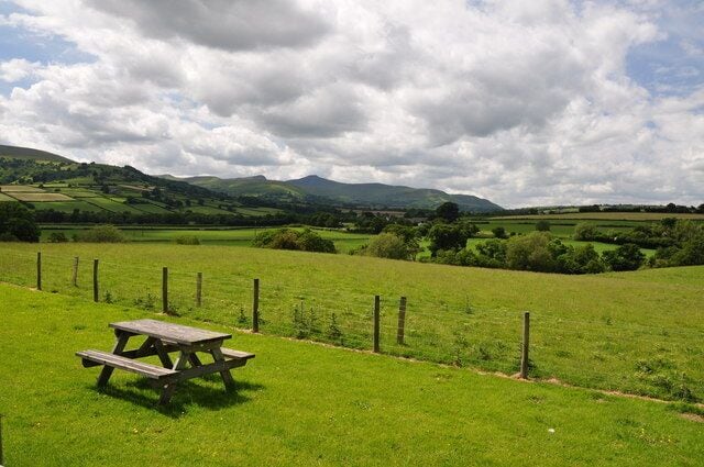 A picnic table in a grassy field with a fence and hills in the background.