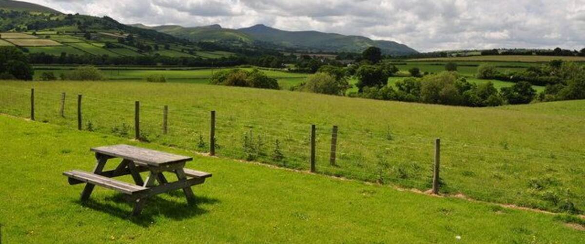 A picnic table in a grassy field with a fence and hills in the background.