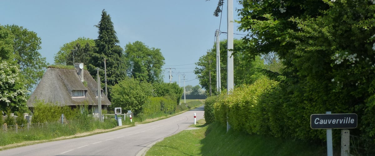 A rural road with a sign for Cauerville, surrounded by greenery and a thatched-roof house.