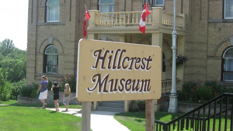 A sign for Hillcrest Museum with a brick building in the background.