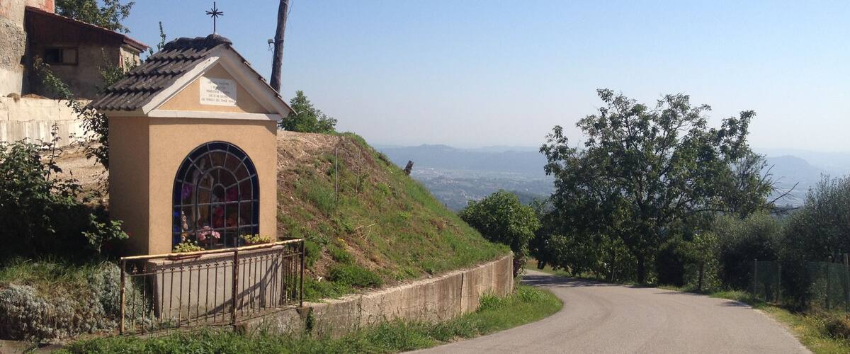 A rural road curving through a hilly landscape with a small chapel on the left and a view of distant mountains.