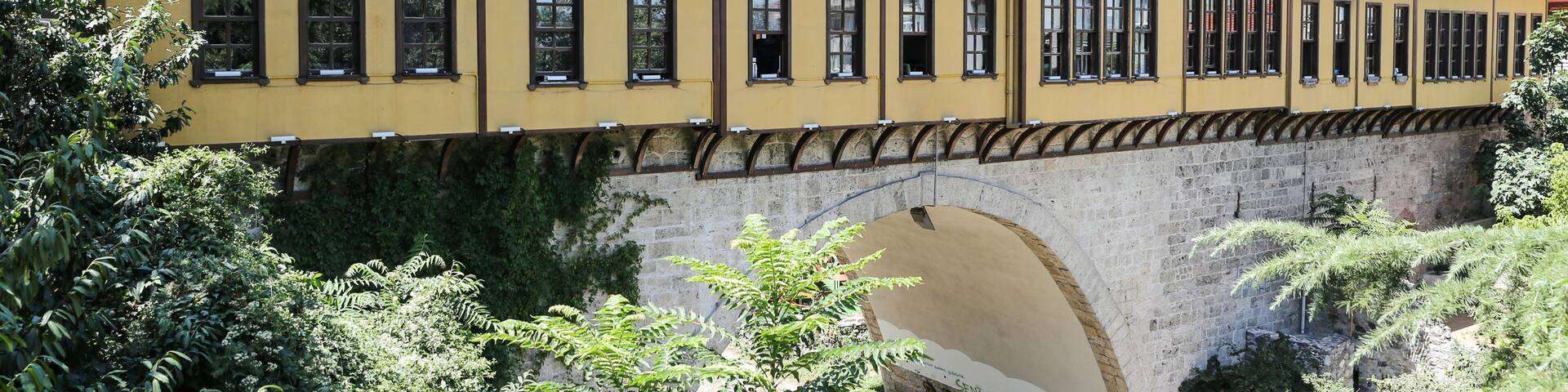 A yellow building with a red-tiled roof, a stone bridge, and surrounding greenery.