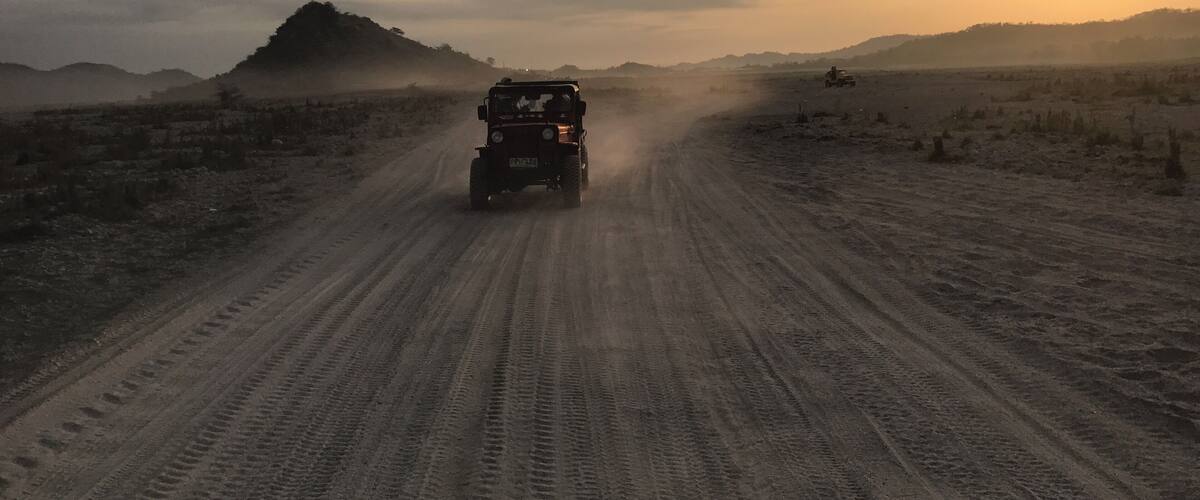 A jeep driving on a dusty road with a mountain in the background and the sun setting.