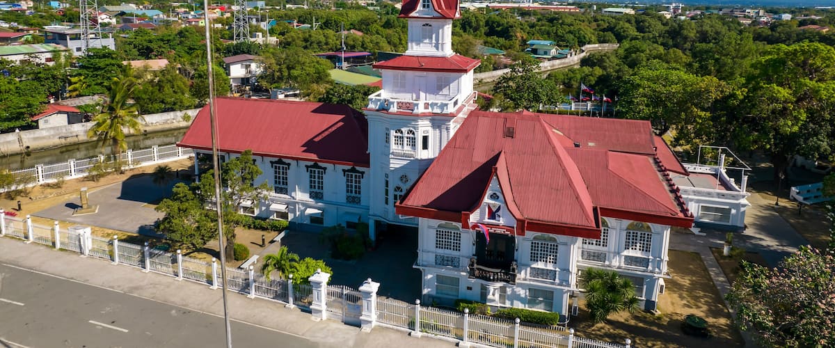 A white building with a red roof and a tower, surrounded by greenery and a river.