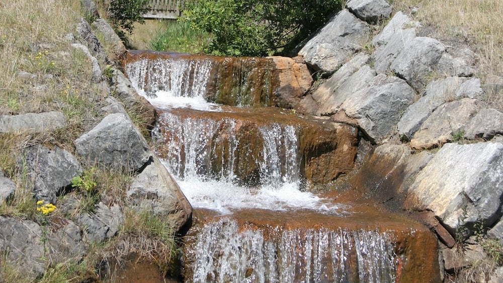Une petite cascade se déversant sur des rochers, dans un cadre naturel, avec une colline herbeuse et un arbre en arrière-plan.
