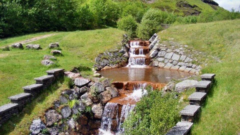 Una pequeña cascada que se precipita a un estanque rodeado de escalones de piedra y abundante vegetación.