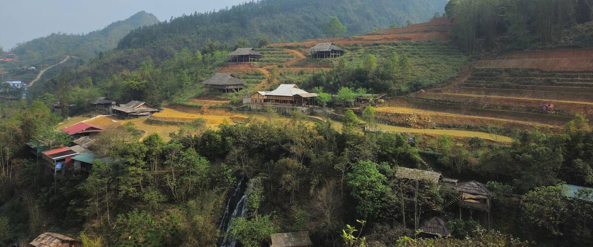 Terraced rice fields, traditional houses, and a waterfall in a hilly landscape.
