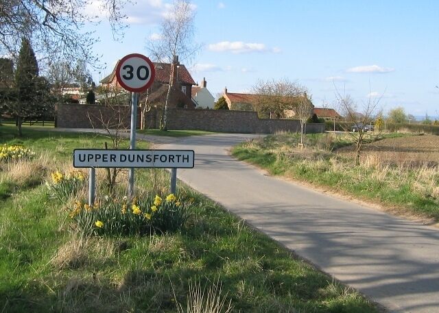 A rural road with a speed limit sign of 30, surrounded by fields and houses.