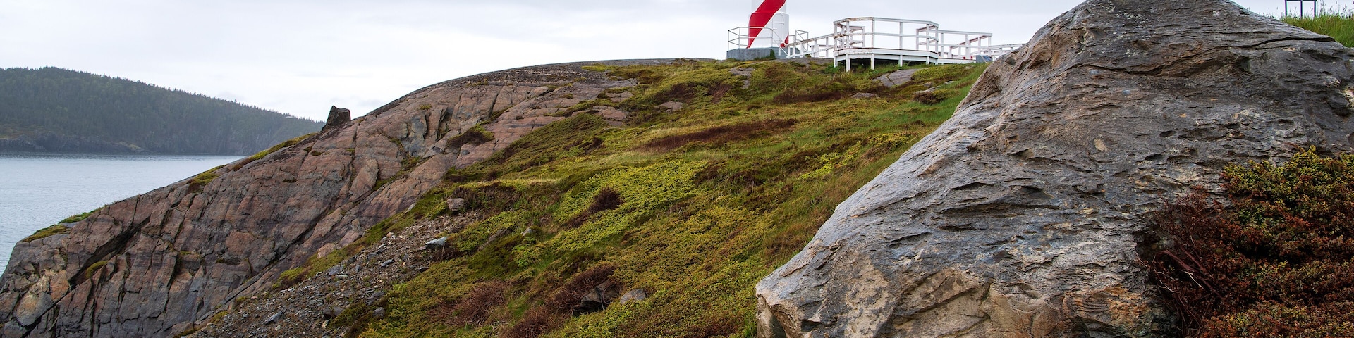 Um farol numa falésia rochosa à beira-mar, rodeado por vegetação e um céu nublado.