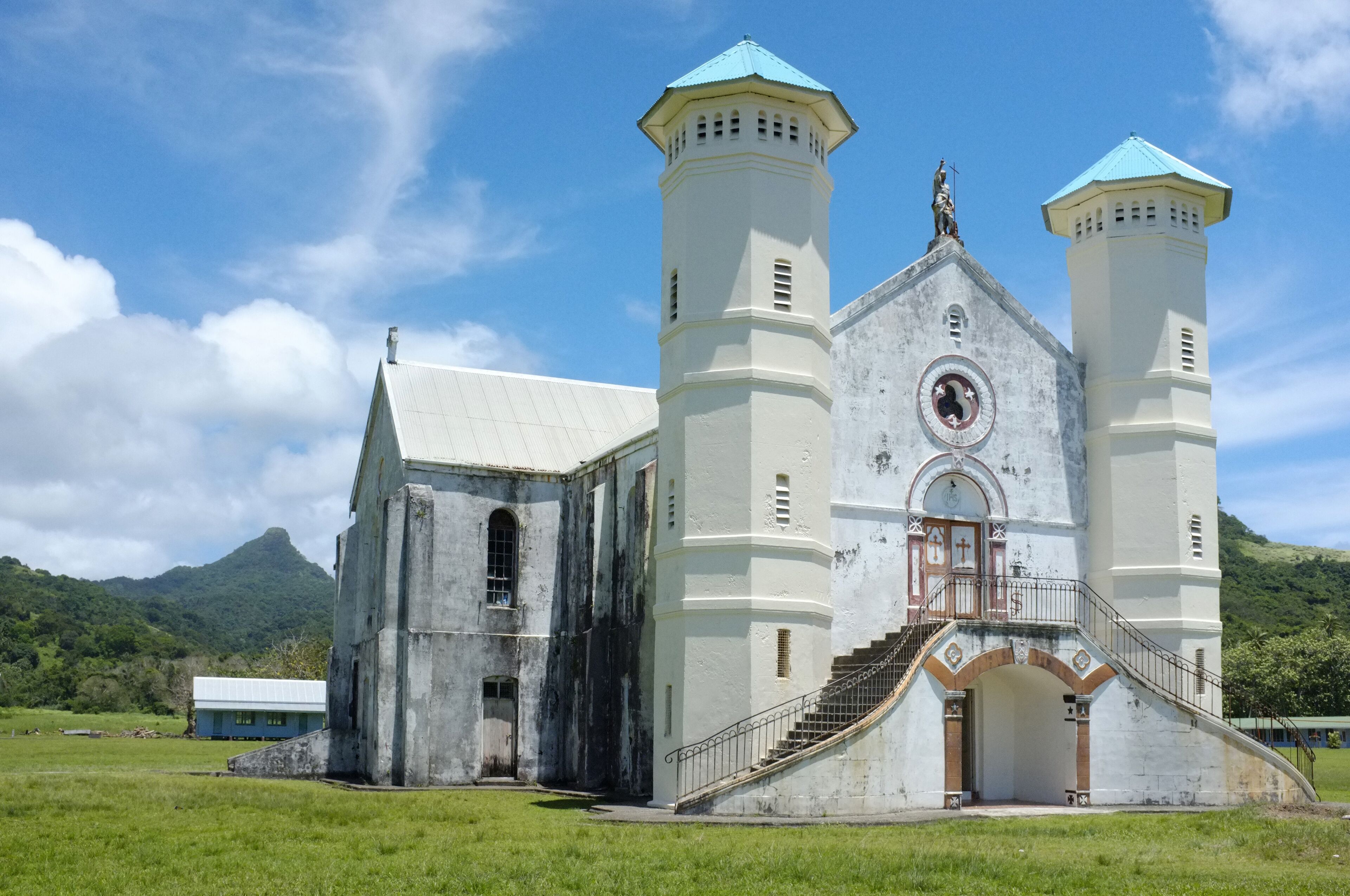 A white church with two towers and a statue on top, surrounded by green fields and mountains.