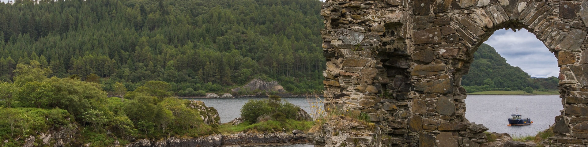 Stromeferry, Scotland - June 10, 2012: Window in rock wall of Castle Strome ruins on green hill. Cloudscape and Loch Carron with motor boat seen in back. Mountains on opposite shore and horizon.
