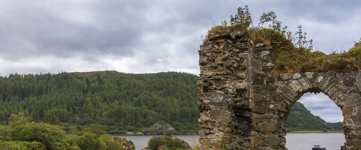 A stone arch ruin by a lake with a boat in the distance and a forested hill in the background.