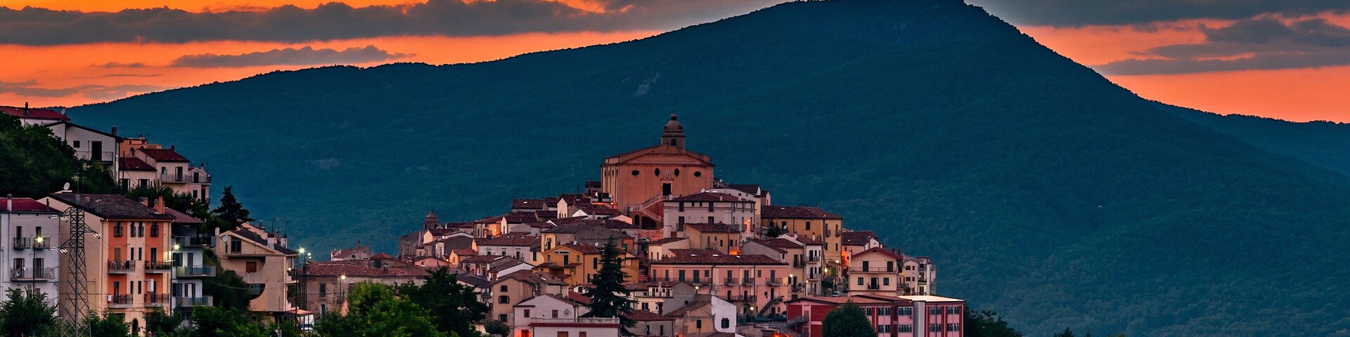 Un pueblo costero con edificios coloridos y una iglesia destacada, con vistas a una montaña y un cielo al atardecer.