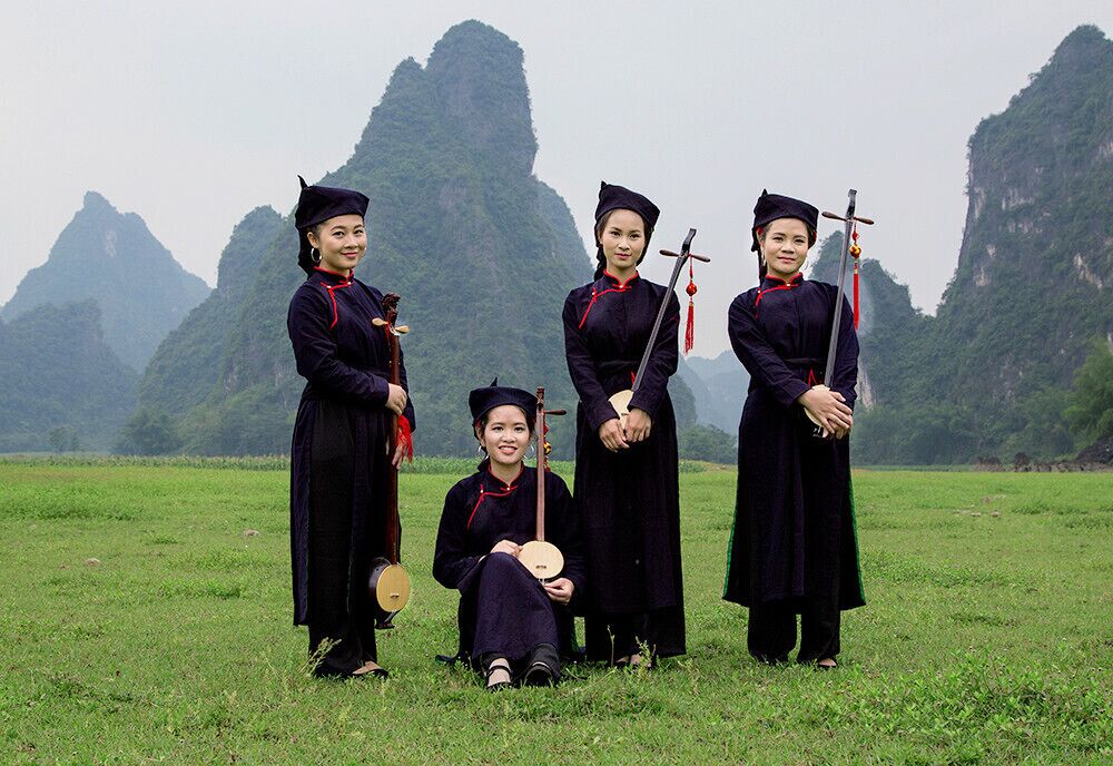 A group of people in traditional attire standing in a grassy field with mountainous terrain in the background.
