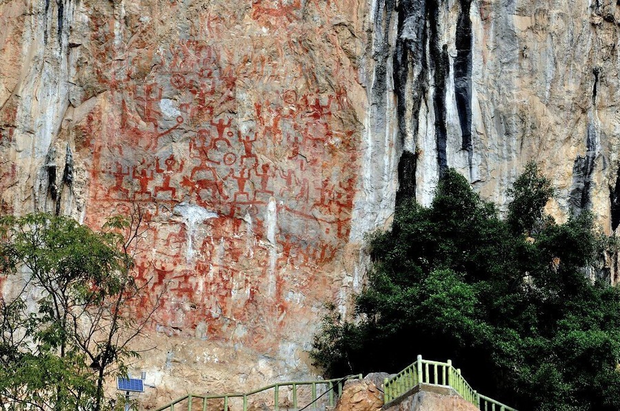 Una pared de roca con antiguas tallas, una escalera y vegetación circundante.