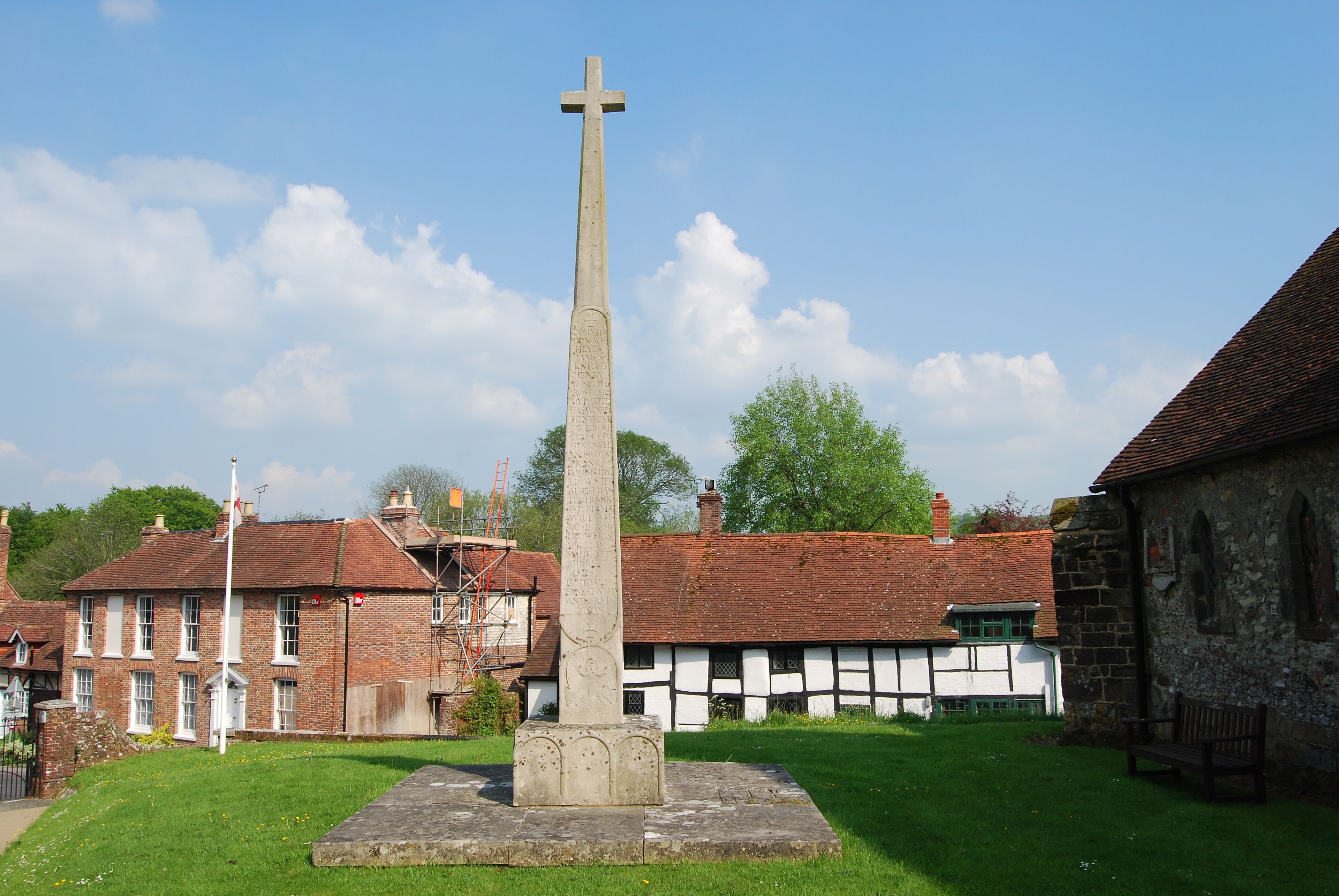 A tall stone cross in a grassy area with traditional buildings in the background.