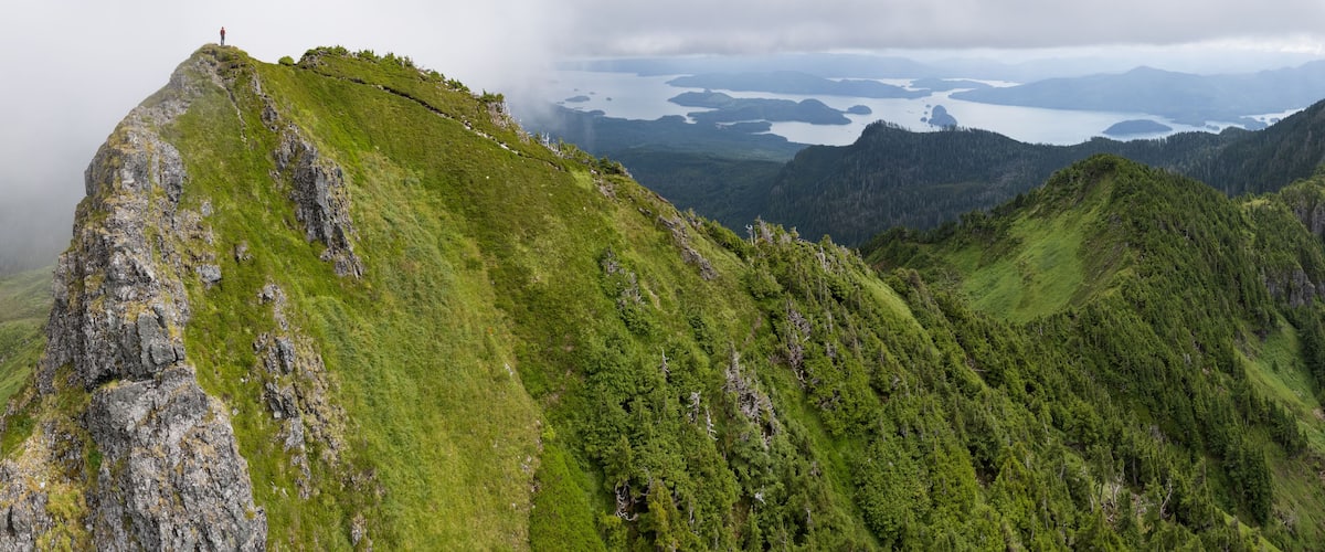 A mountain peak with a person standing at the top, surrounded by lush green forests and a lake in the background.
