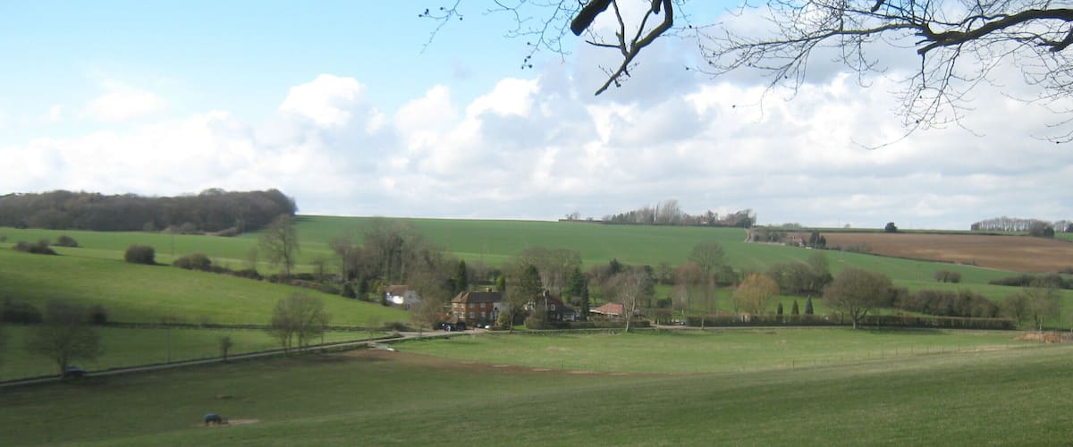 A rural landscape with rolling green hills, scattered trees, and a few buildings in the distance.