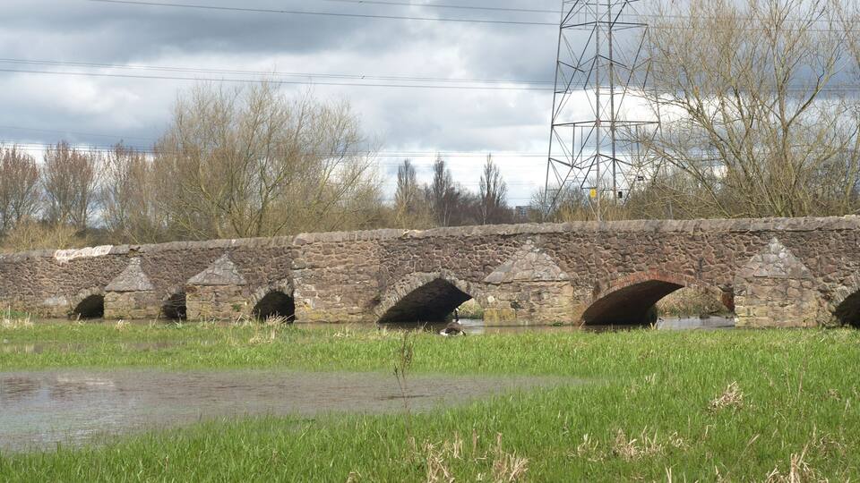 A stone bridge with multiple arches over a river, surrounded by grass and trees.