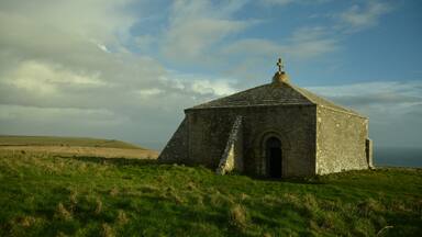 A stone building with a cross on top, surrounded by green grass and a cloudy sky.