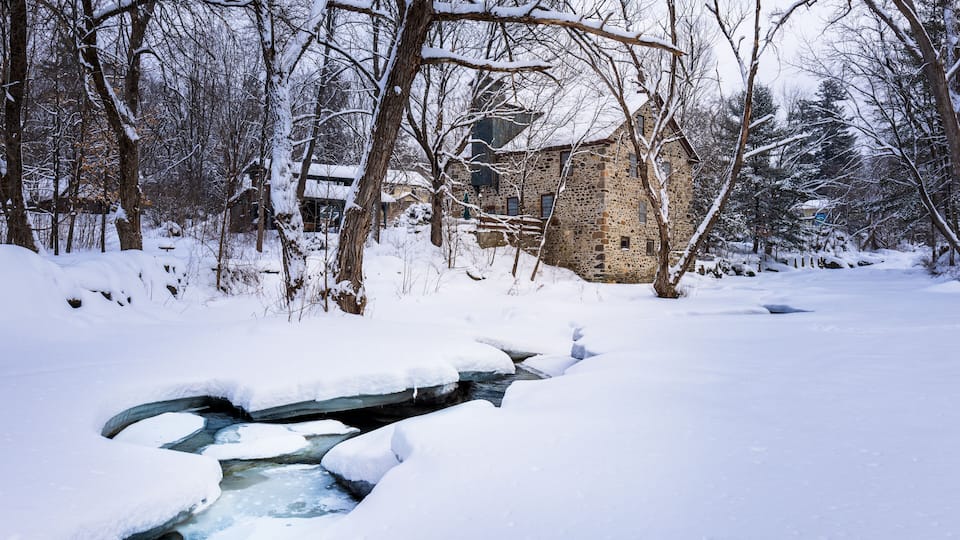 A stone house surrounded by snow-covered trees and a partially frozen stream.