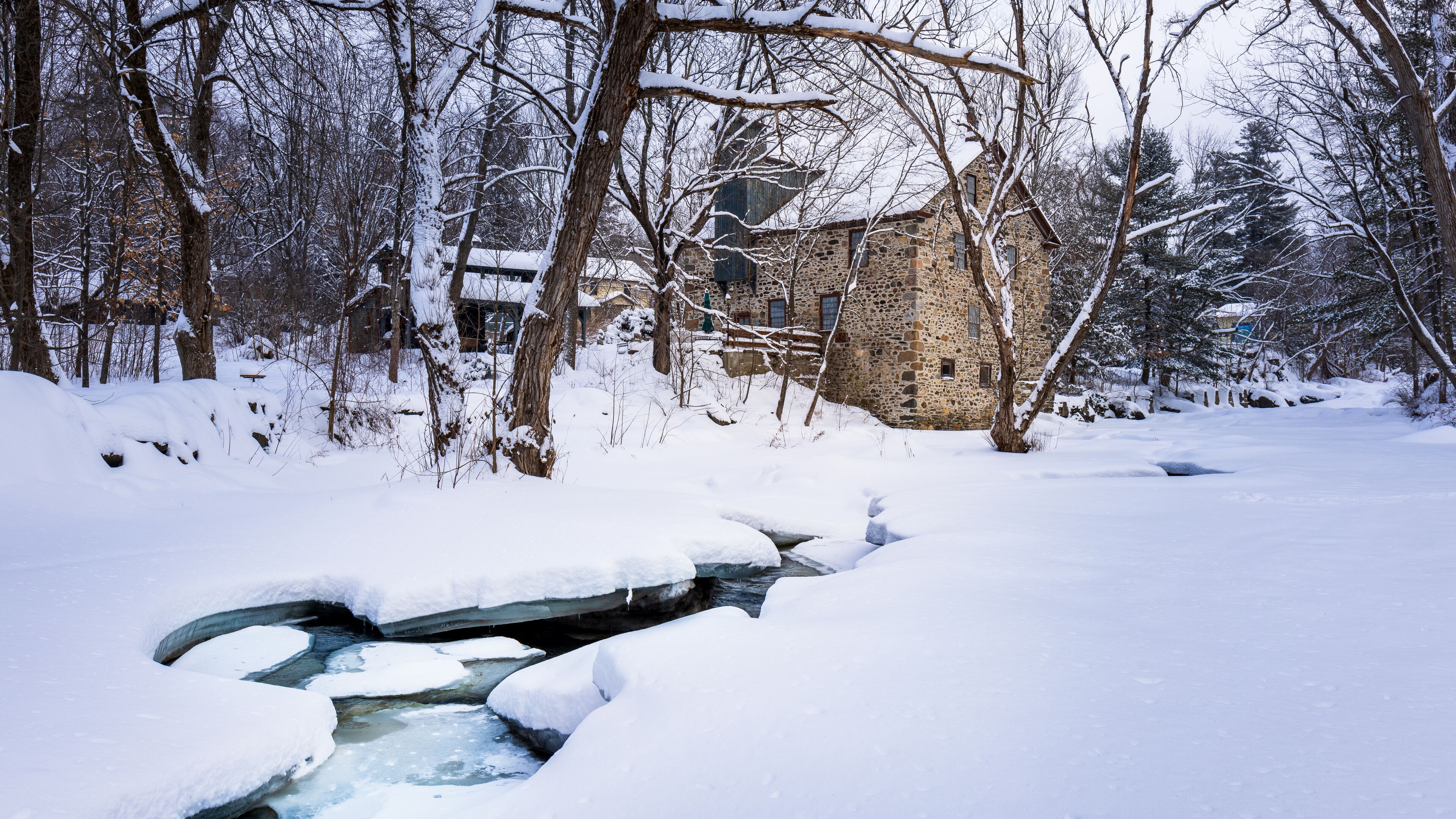 A stone house surrounded by snow-covered trees and a partially frozen stream.