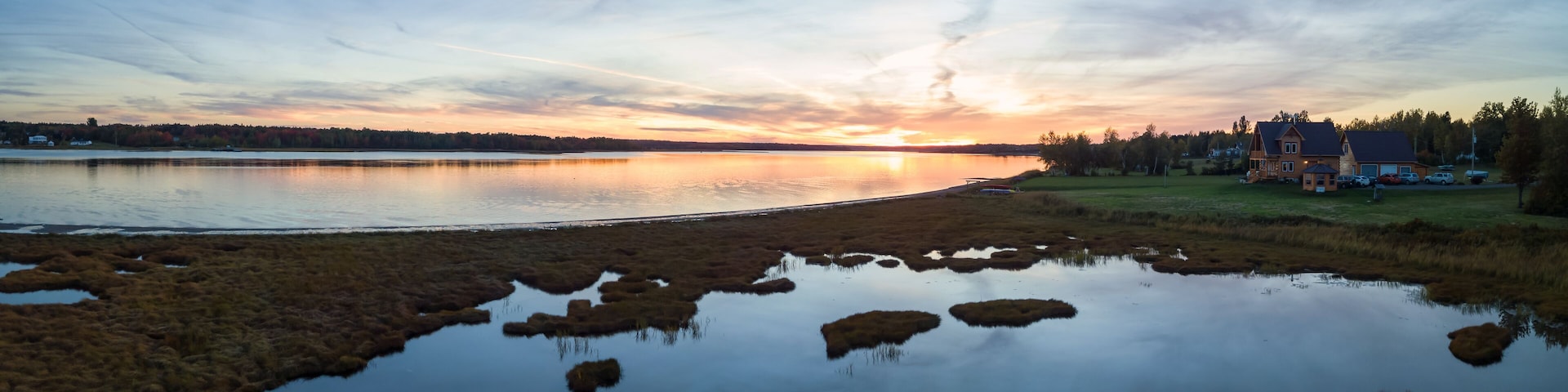 Um pôr do sol sobre um lago calmo, com reflexos do céu, cercado por áreas gramadas e uma área residencial com casas.