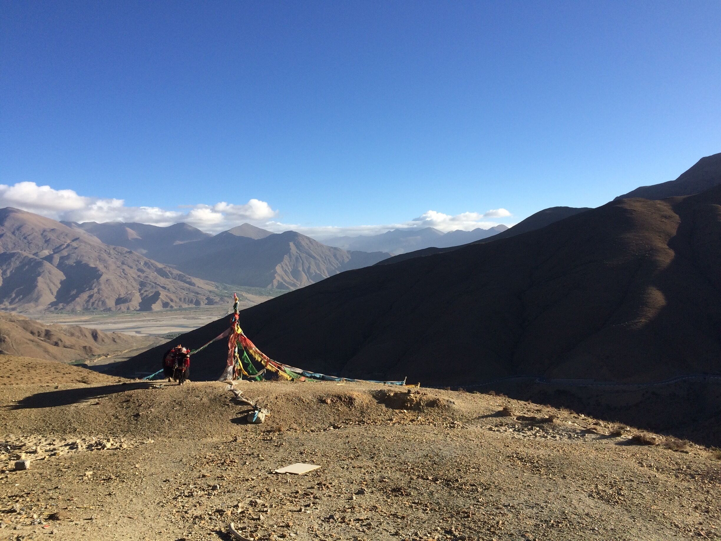 A mountainous landscape with a clear sky, a small shrine with colorful prayer flags, and distant mountains.