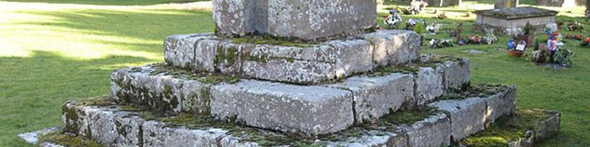Um monumento de pedra com degraus e uma cruz, rodeado por um cemitério com lápides.