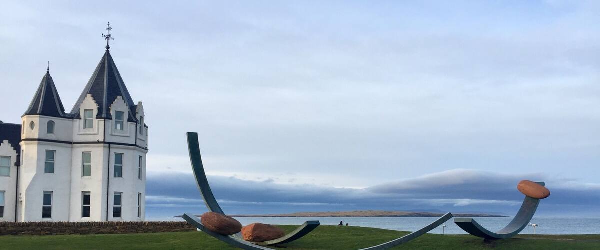 A castle with a white facade and black roof, a grassy area with a large metal sculpture, and a body of water in the background.