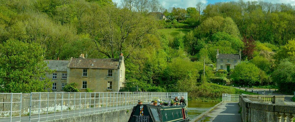 A narrow canal with a narrowboat docked at the side, surrounded by lush greenery and hills.