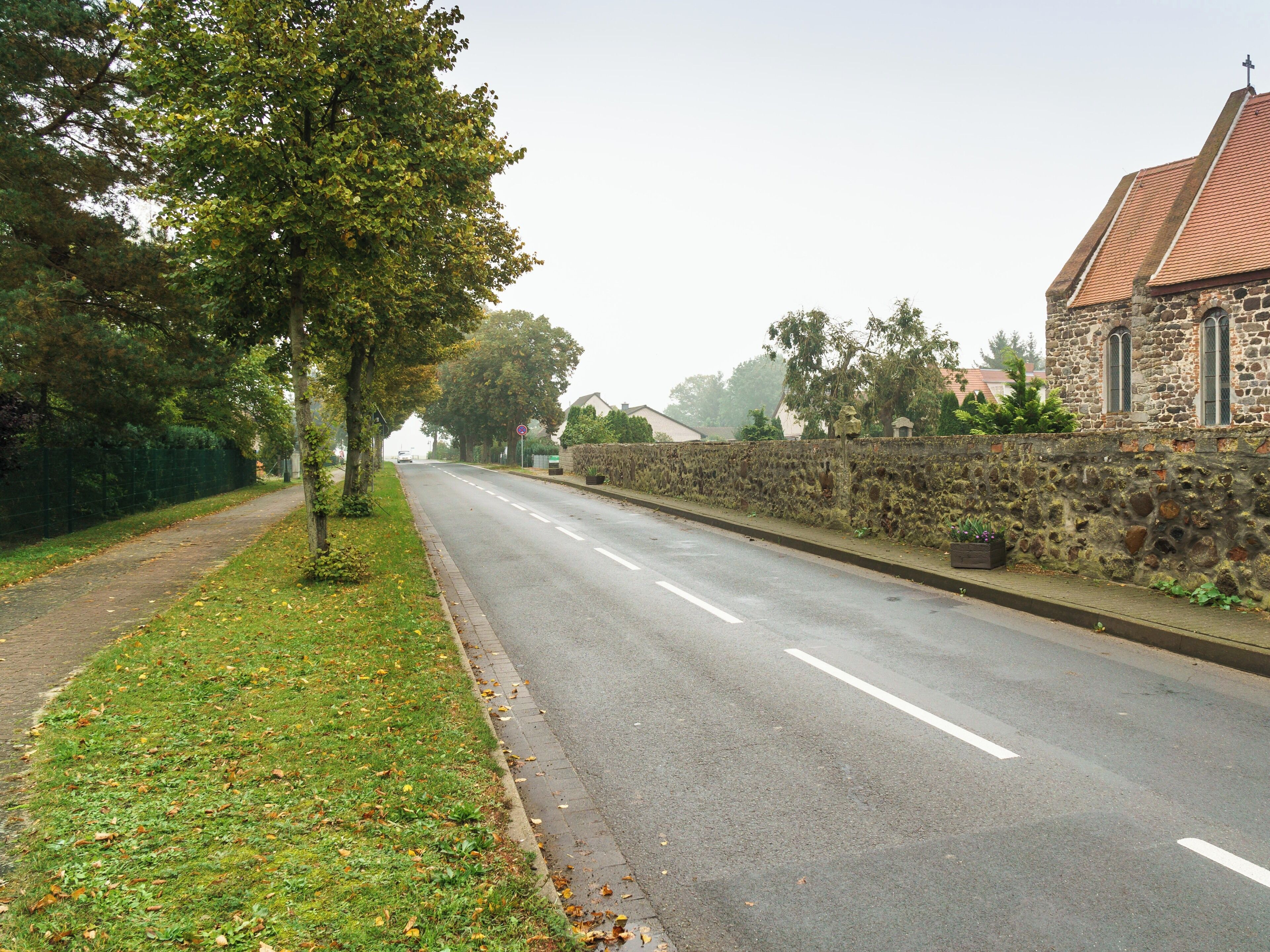 A quiet street lined with trees and a stone wall, leading to a stone building with a cross on top.