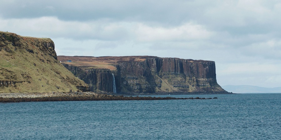 A coastal cliff with a waterfall, a rocky shoreline, and a calm sea under a cloudy sky.