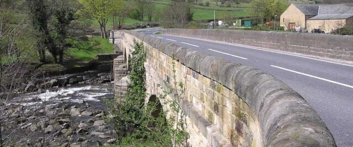 A stone bridge over a river, a road, and rural buildings in the background.