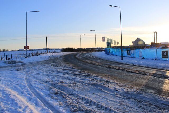 Uma estrada coberta de neve com sulcos de pneus, uma cerca e um edifício ao fundo.