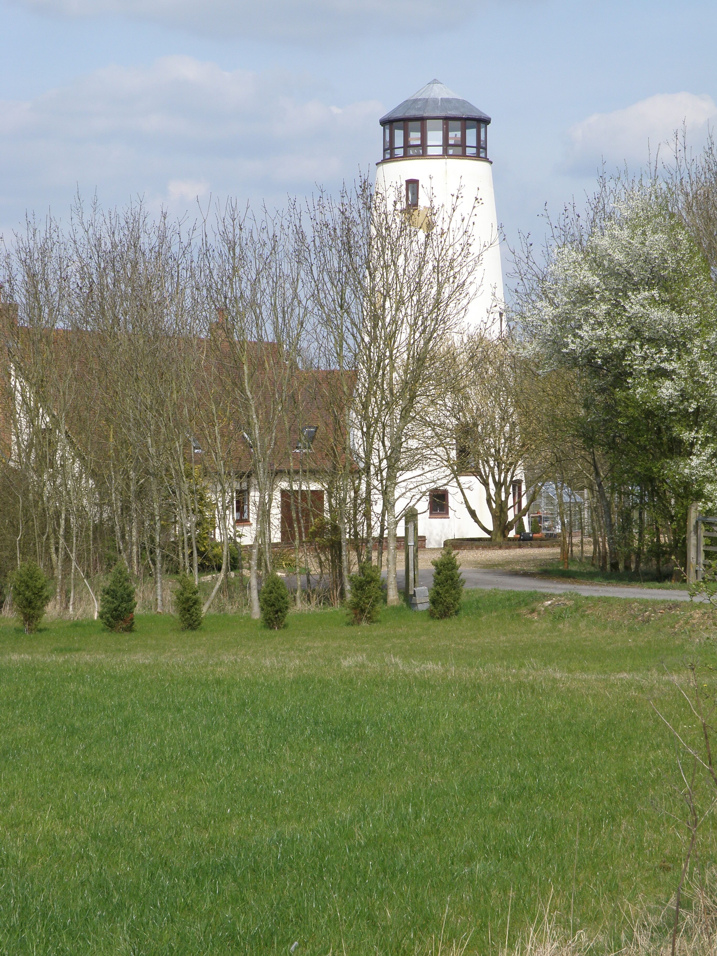 A white tower with a dark roof, surrounded by trees and a grassy field.
