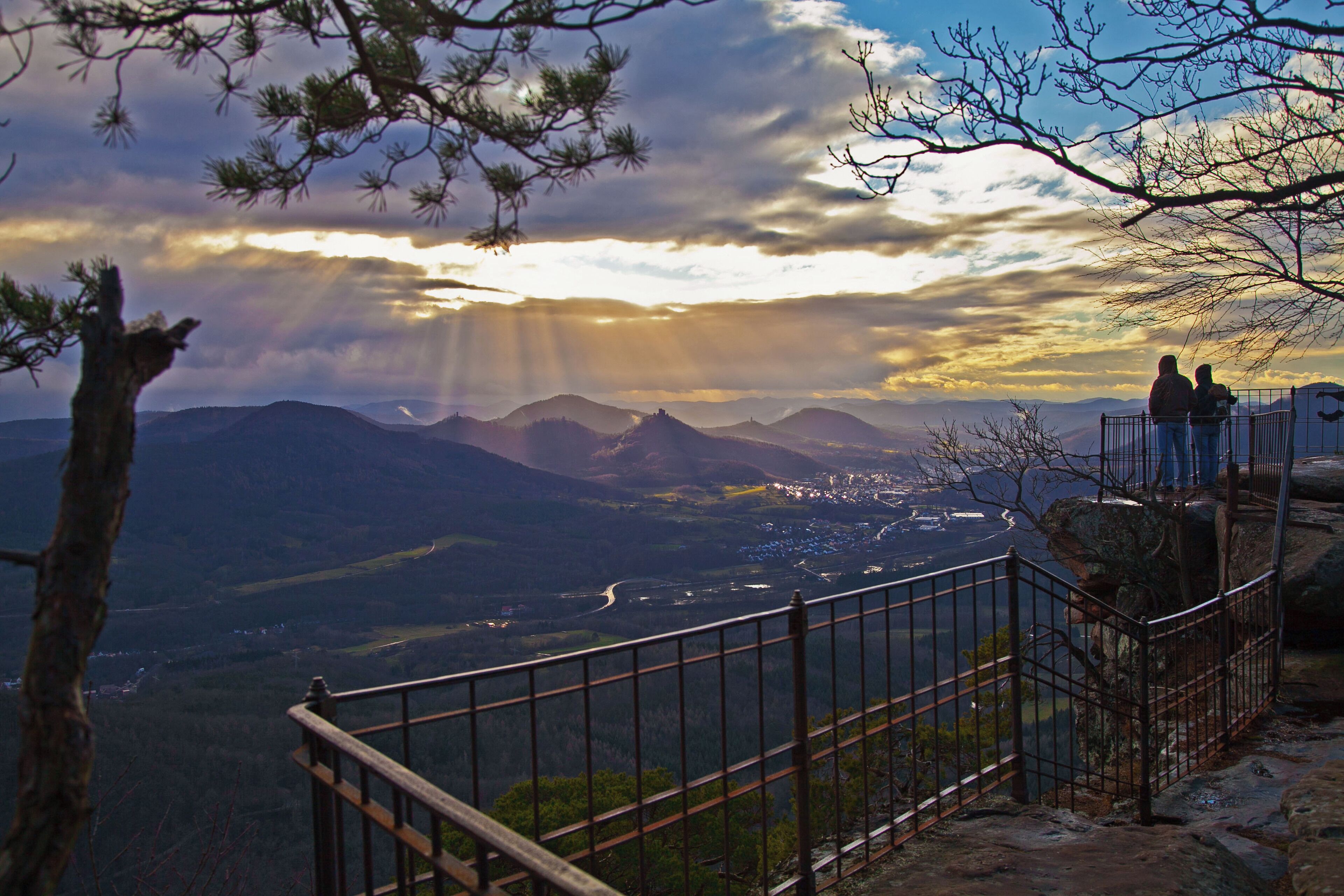 A mountainous landscape with a sunset, a railing, and two people overlooking the view.