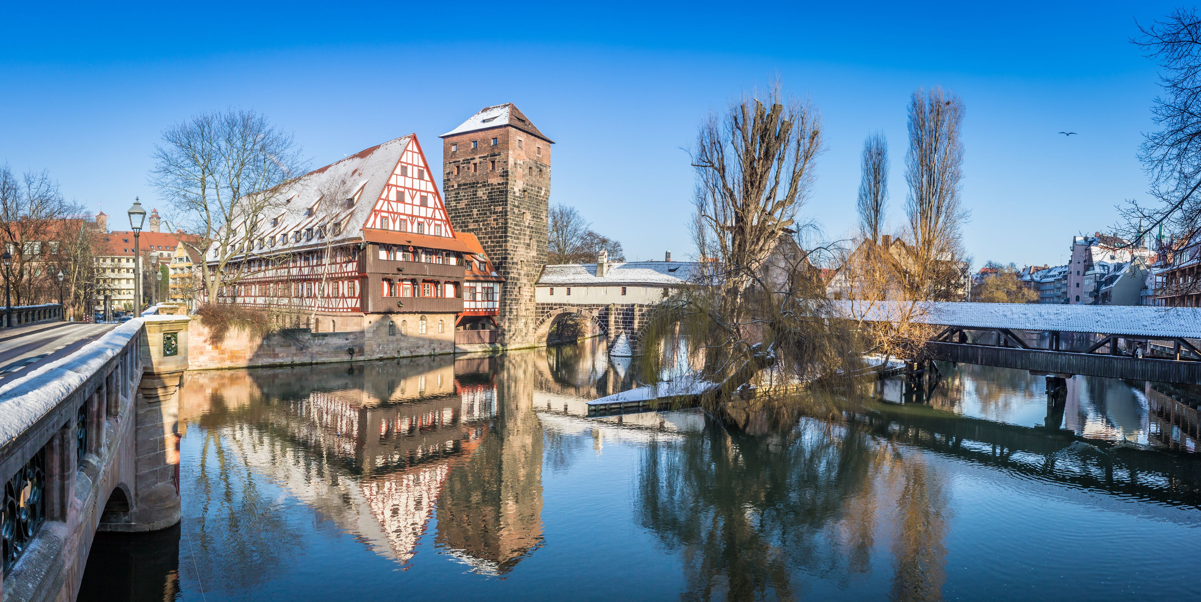 Un río que refleja edificios, un puente y árboles desprovistos de hojas bajo un cielo despejado.