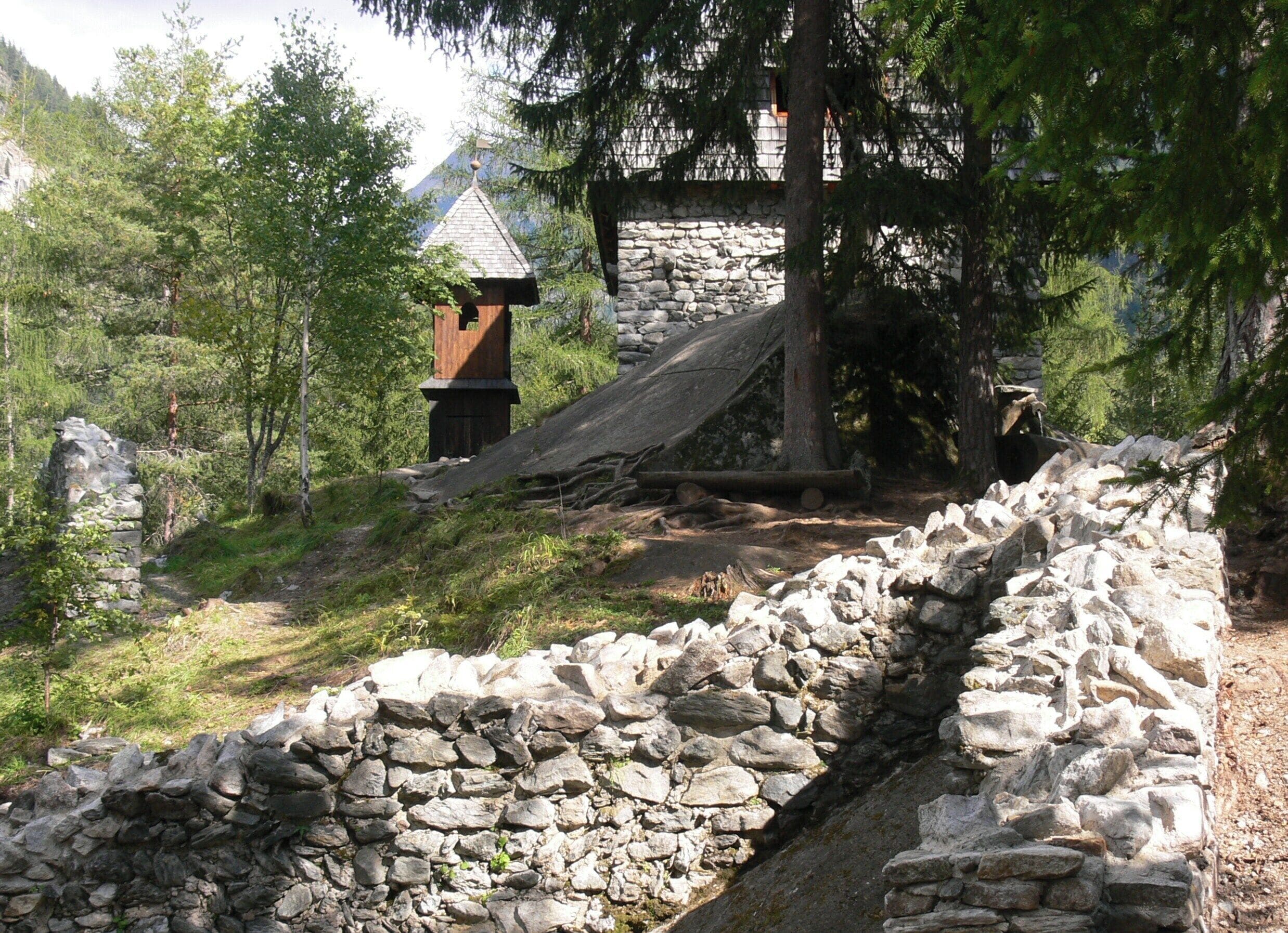 A stone wall, a wooden structure, and a forested area with a mountain in the background.