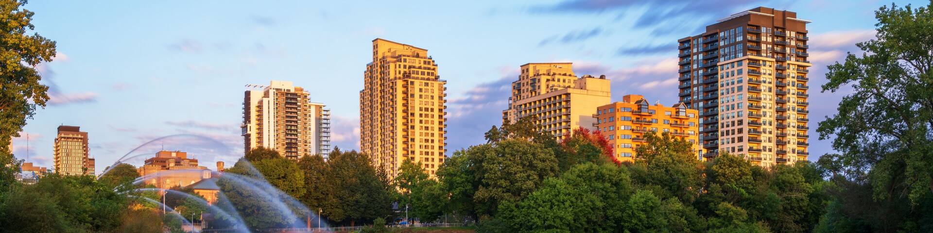 A calm lake with a fountain in the foreground, surrounded by lush greenery and tall buildings in the background.