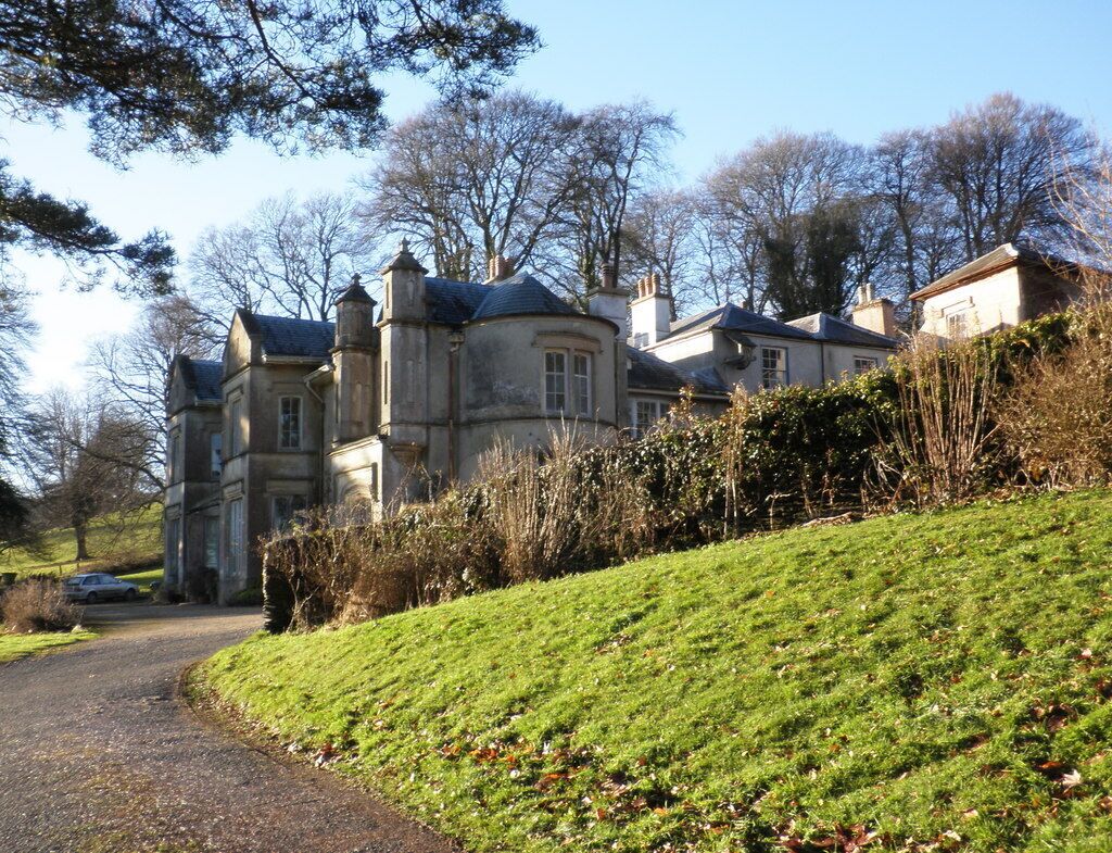 A large, historic stone house with a sloped roof, surrounded by a well-maintained lawn and trees.