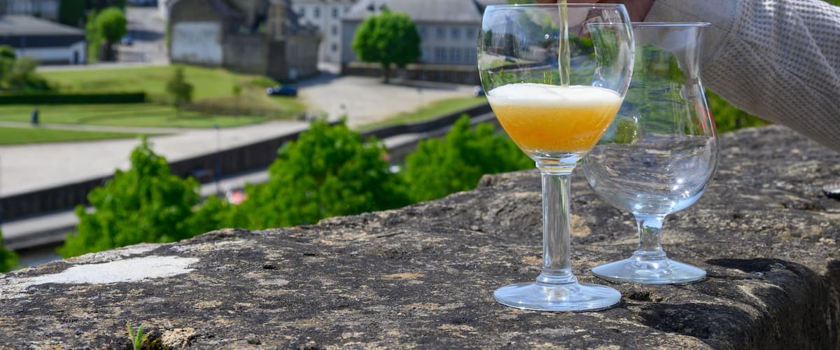 A hand pouring a drink into a glass with a scenic view of buildings and greenery in the background.