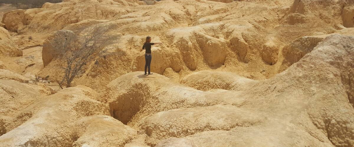 A person standing in a dry, rocky landscape with eroded hills and sparse vegetation.